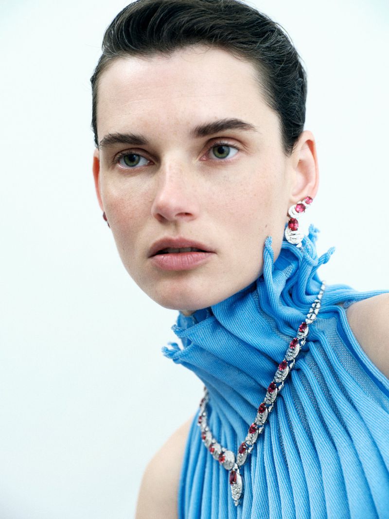 Close-up of a woman with short dark hair wearing red and white earrings and necklace, and a blue textured top against white background.