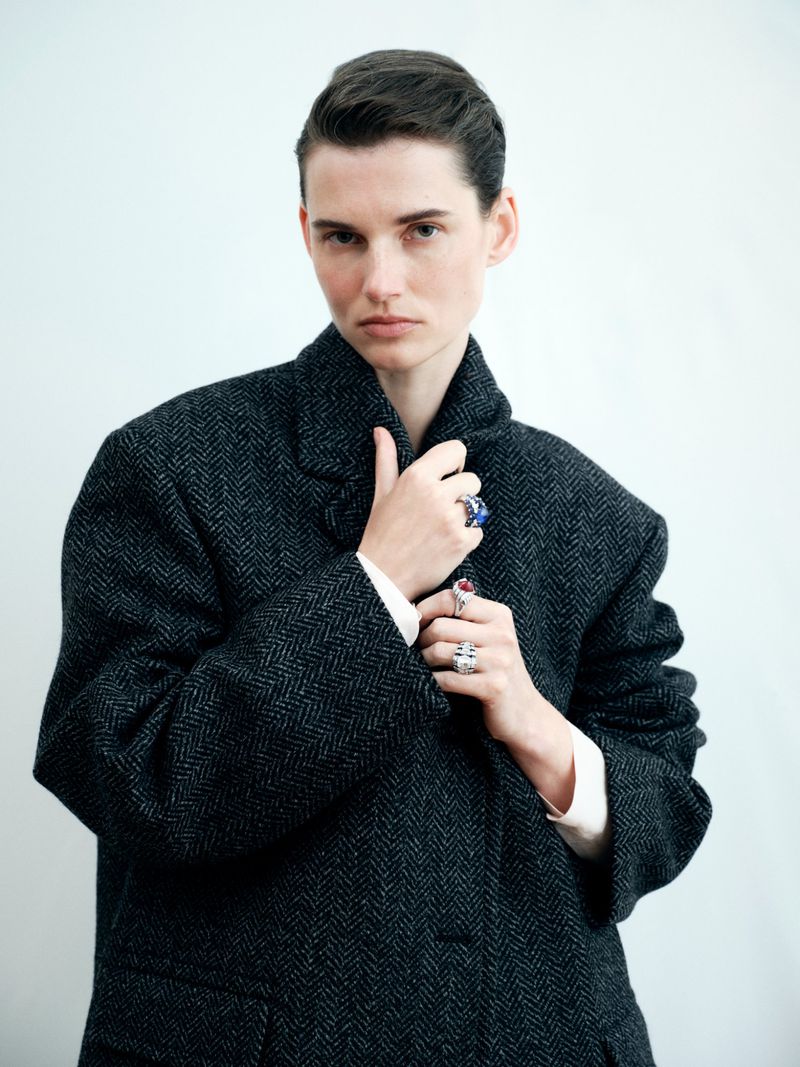 Portrait of a woman with short hair wearing a dark grey herringbone coat with jewelry against a white background in studio lighting.