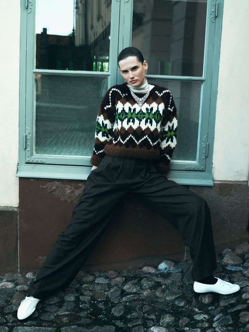 An adult woman poses against a light blue window wearing a patterned knit sweater, dark pants, and white sneakers on a stone walkway.
