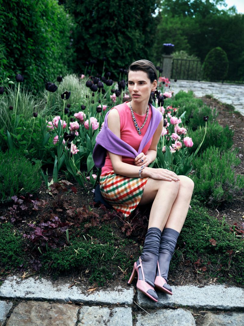 A woman in a pink top and plaid skirt sits in a garden among pink and purple tulips, wearing gray socks and pink heels, with trees in the background.