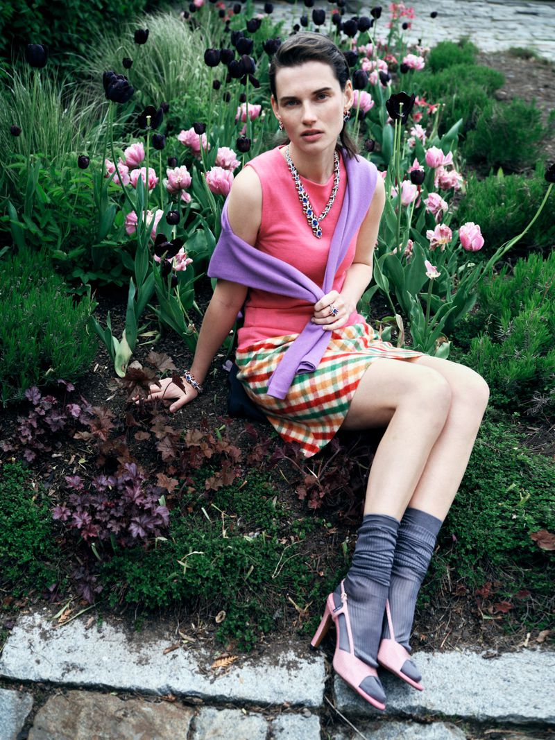 Adult female model sits in a lush outdoor garden wearing pink top, plaid skirt, purple sweater, necklace and pink high heels in daylight.