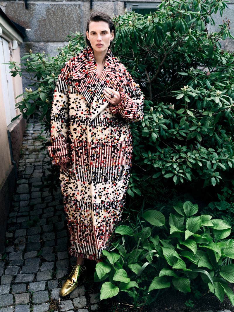 A stylish adult woman is posing in an outdoor setting, wearing a patterned coat and gold shoes, standing near green plants and a stone building.
