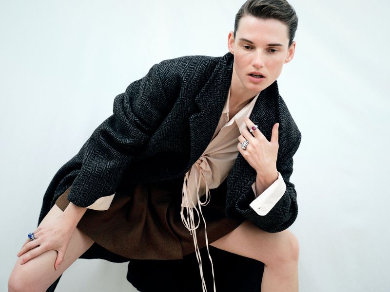 A model poses in a studio wearing a grey jacket, brown skirt, and light pink blouse. The image is shot in portrait format against a white backdrop.