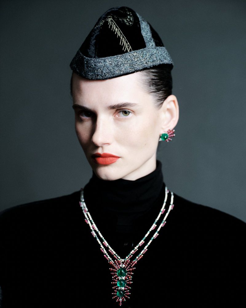 Elegant woman in a black turtleneck, jeweled necklace and earrings, and a patterned cap against a neutral gray studio backdrop. 