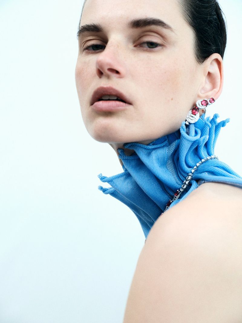 Close-up fashion shot of a woman with light skin and short brown hair wearing artistic jewelry and a unique blue ruffled collar.