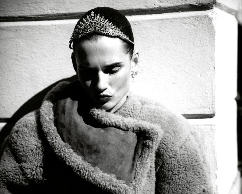 Black and white portrait of a woman wearing a jeweled headband and shearling coat, gazing downward in an outdoor setting with natural light.