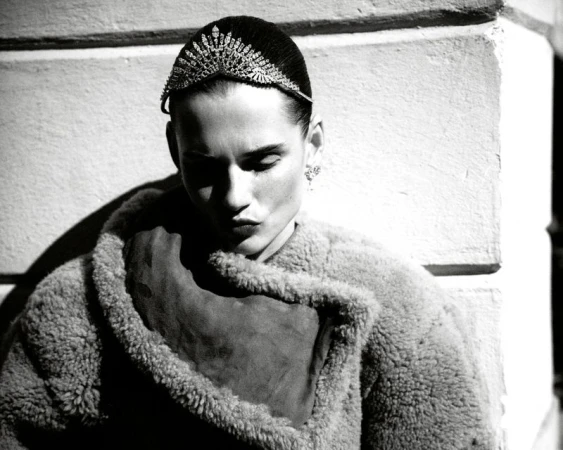 Black and white portrait of a woman wearing a jeweled headband and shearling coat, gazing downward in an outdoor setting with natural light.