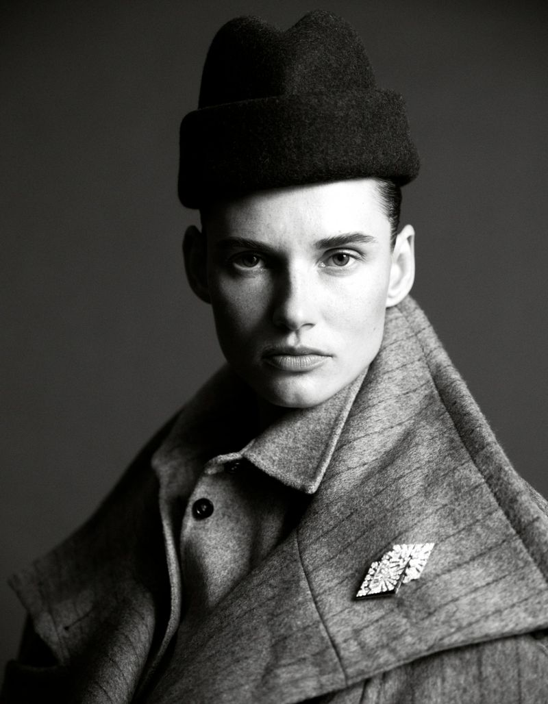 Black and white portrait of a fashionably dressed person wearing a hat and brooch, posing in front of a neutral background.