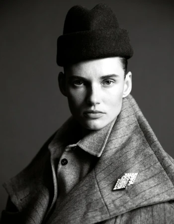Black and white portrait of a fashionably dressed person wearing a hat and brooch, posing in front of a neutral background.