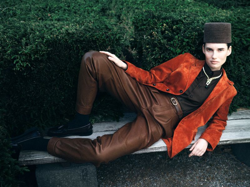 Stylish model with a hat poses on a bench outdoors wearing an orange jacket, brown pants, black shoes and socks, and a gold necklace.