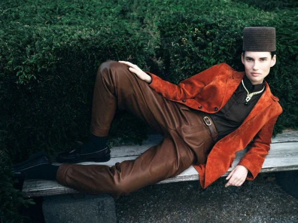 Stylish model with a hat poses on a bench outdoors wearing an orange jacket, brown pants, black shoes and socks, and a gold necklace.