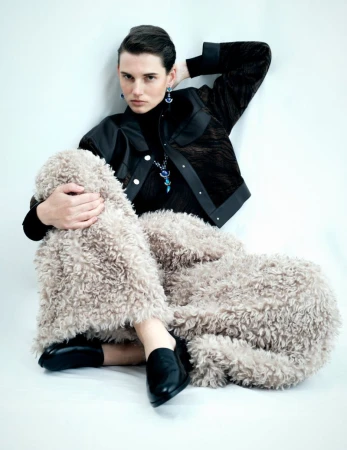 An adult female model is sitting in a black jacket and furry pants in a white photo studio, gazing directly at the camera in a fashion shot.