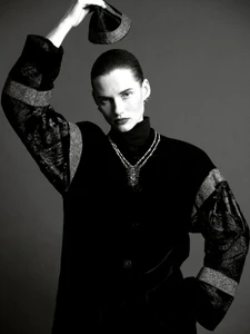 Model Giedre Dukauskaite wearing a dark patterned jacket and statement necklace, holding a small hat above her head in a studio.