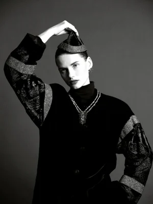 Monochrome fashion portrait of Giedre Dukauskaite wearing a structured jacket, layered necklace, and holding a small hat above her head.