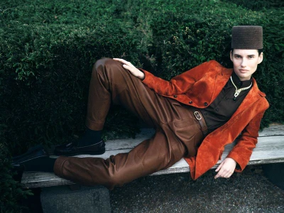 Model Giedre Dukauskaite in rust suede jacket and brown leather trousers reclining on a rustic wooden bench before dense green foliage.