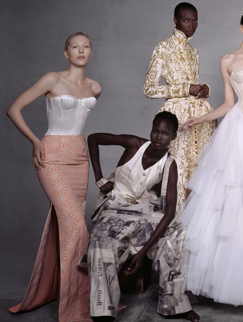 Fashion editorial shot featuring four models in dresses. The models pose in front of a plain grey background in a studio environment.
