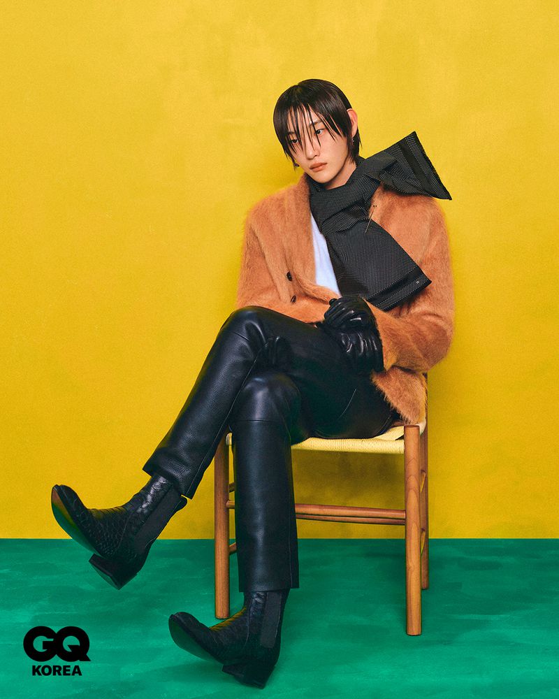 A stylish, young adult with dark hair and bangs poses while sitting on a chair in a brown coat, black scarf, leather pants, and black boots.