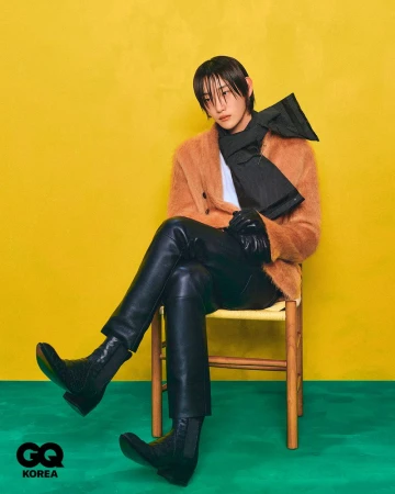 A stylish, young adult with dark hair and bangs poses while sitting on a chair in a brown coat, black scarf, leather pants, and black boots.