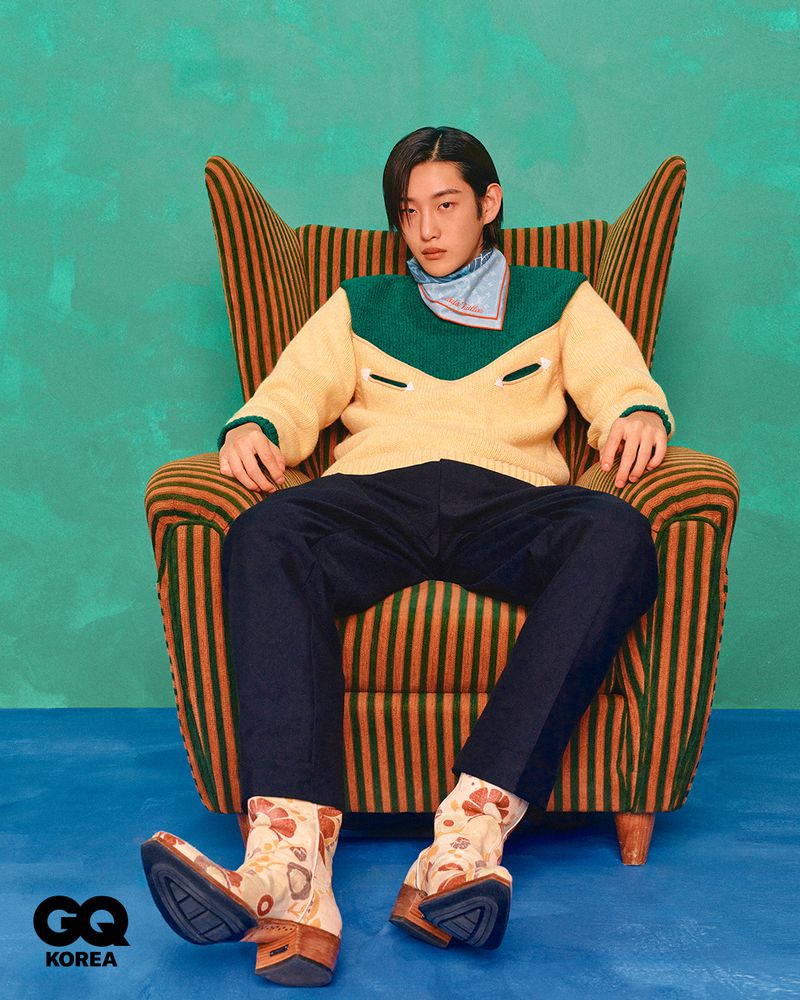 Man sits in a striped armchair, wearing a two-tone sweater, dark pants, and patterned boots in a portrait with a textured green backdrop.