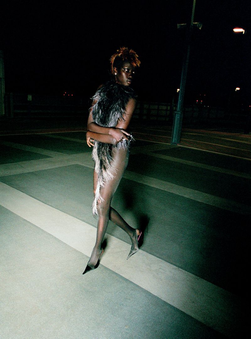 A stylish adult woman is walking outdoors at night, wearing a feathered garment and heels in a dimly lit, urban environment with dark tones.
