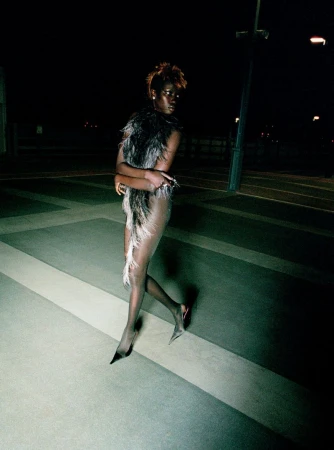 A stylish adult woman is walking outdoors at night, wearing a feathered garment and heels in a dimly lit, urban environment with dark tones.