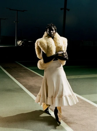 Stylish woman in a faux fur wrap and flowing skirt posing on a tennis court at night, illuminated by artificial light.