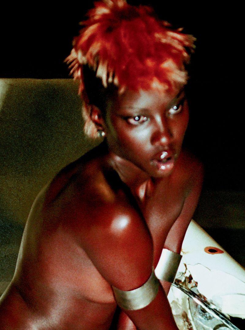 Close-up studio portrait of a young Black woman with reddish-blonde spiky hair, bare shoulders, and reflective silver arm cuff against a dark background.