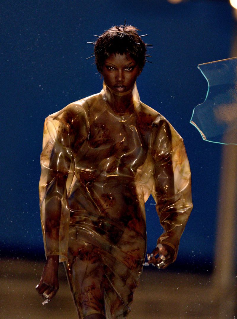 A dark-skinned fashion model showcases a runway look featuring a clear, patterned overcoat and spiky hair accents against a blue backdrop.