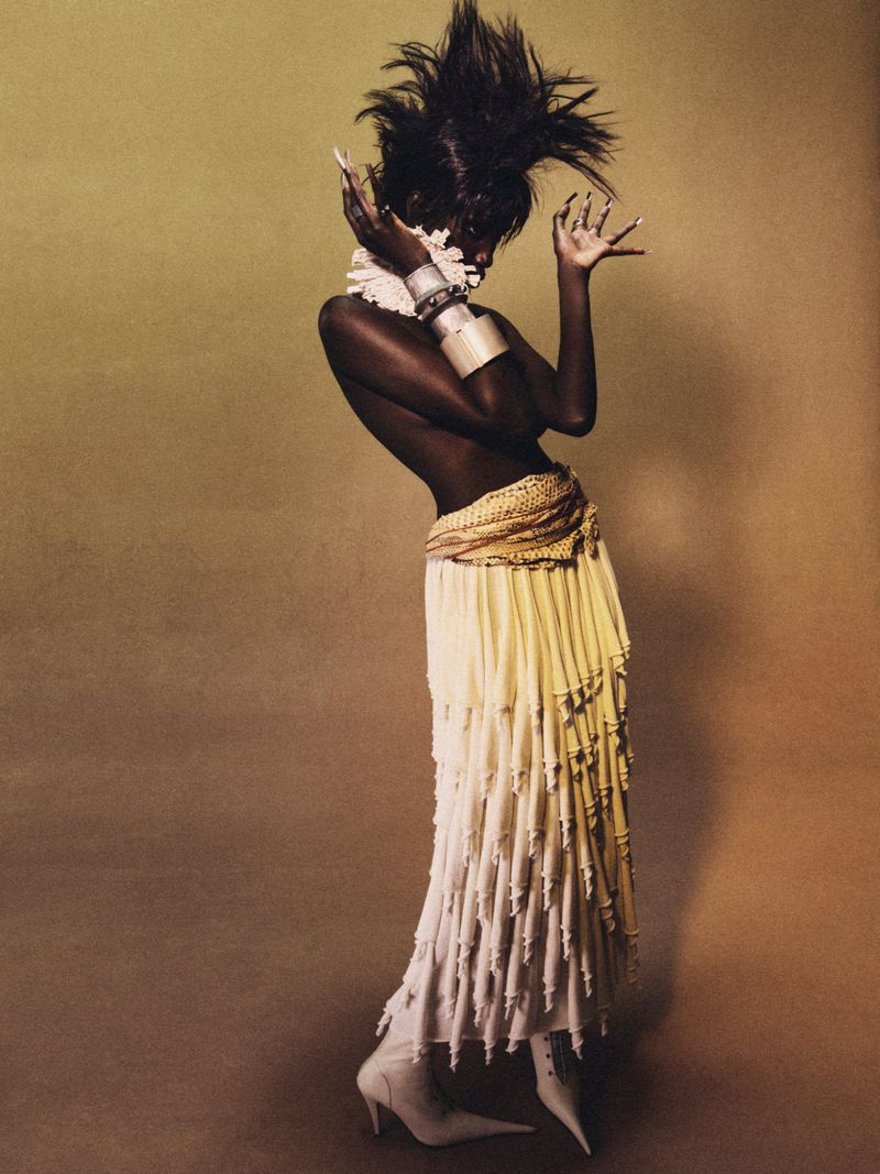A high-fashion portrait of an adult female fashion model posing in a long skirt with fringe, bangles, and high heels against a brown background.