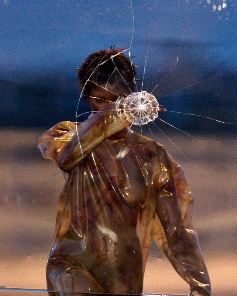 Artistic portrait of a person in a plastic garment, with a shattered glass pane in front, creating a conceptual visual effect.