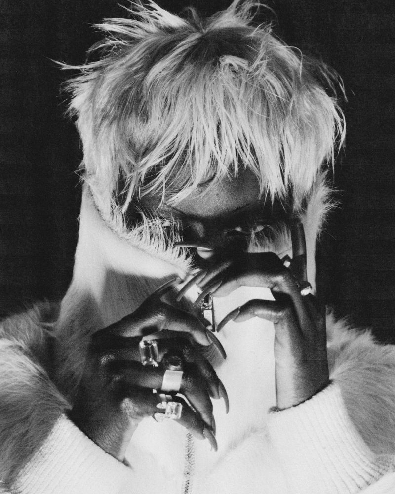 Close-up black and white portrait of a person with blonde tousled hair, rings on their fingers, and a fur collar.