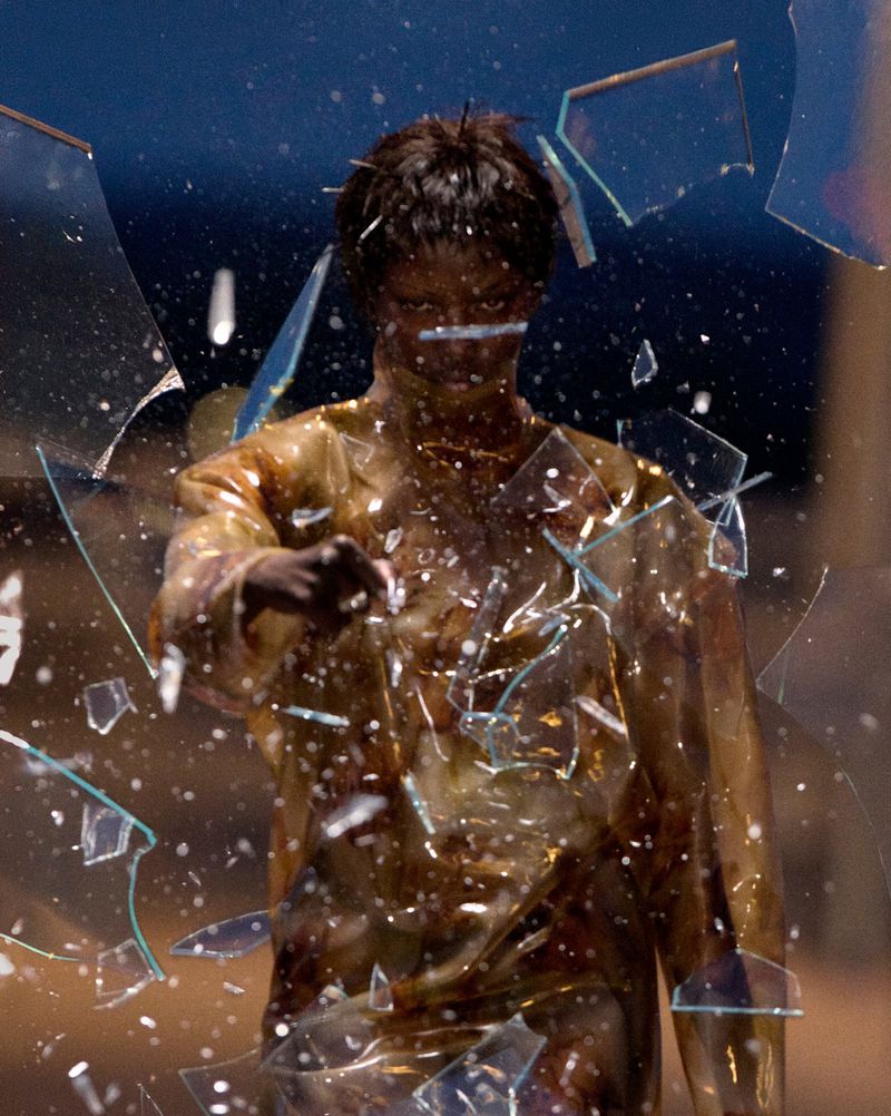 Person standing amidst shattered glass, indoors, with dark blue background, wearing a transparent shirt with gold and brown tones.