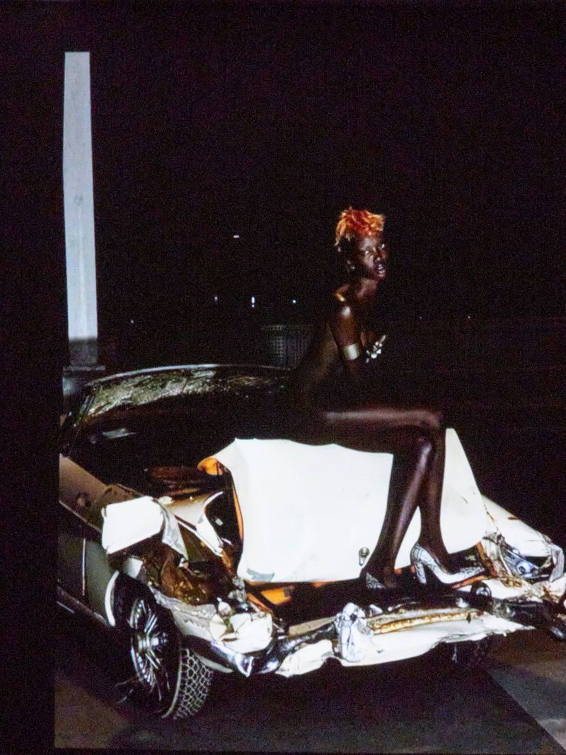 A woman with an orange hairstyle sits on the damaged trunk of a silver car against a dark background, wearing silver high heels.