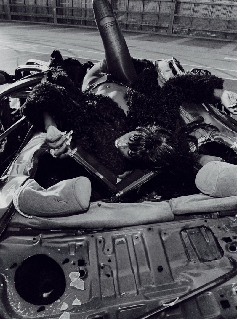 Dramatic black-and-white fashion photo of a Black woman reclining on the wreckage of a car, wearing a dark furry coat.