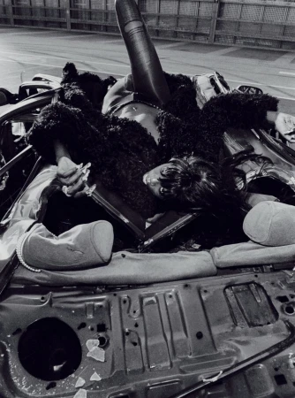 Dramatic black-and-white fashion photo of a Black woman reclining on the wreckage of a car, wearing a dark furry coat.