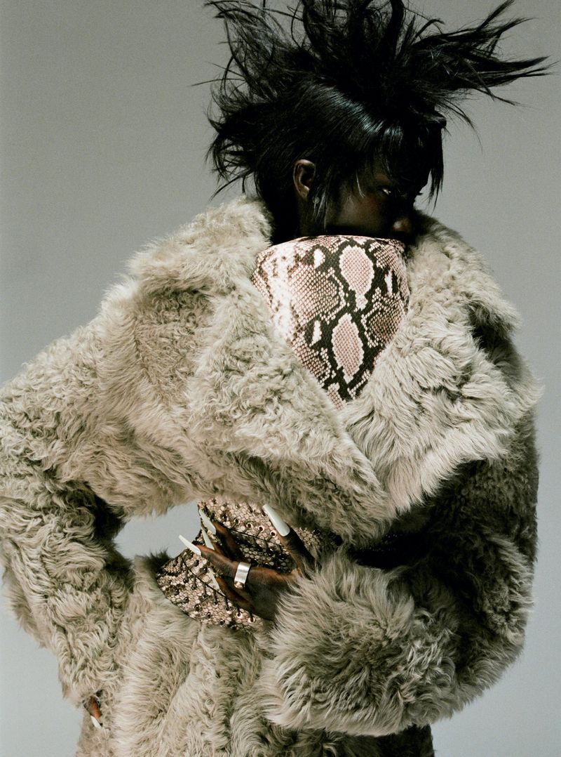 Stylish woman with a spiky hairstyle wearing a large fur coat and carrying a snakeskin print bag in a studio fashion shot.