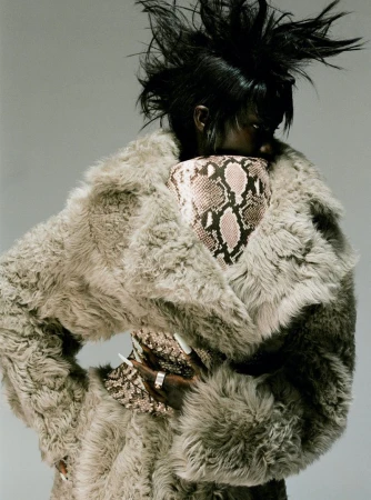 Stylish woman with a spiky hairstyle wearing a large fur coat and carrying a snakeskin print bag in a studio fashion shot.