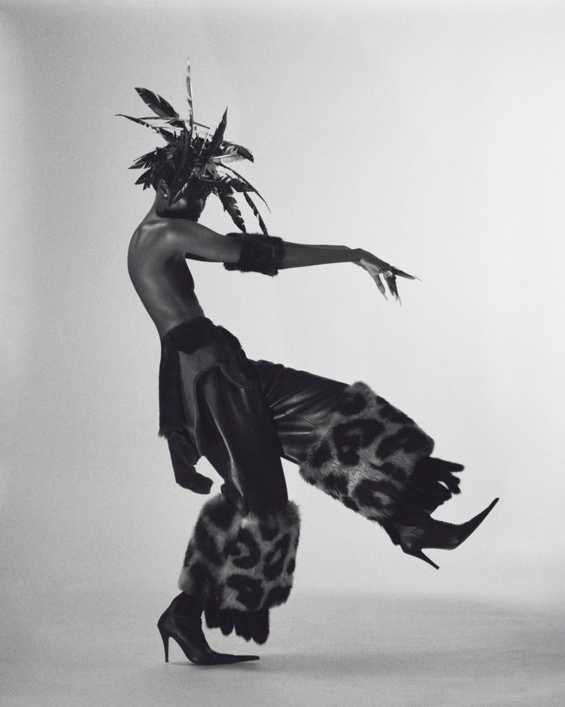 Person in dramatic pose wearing feathered headpiece, animal-print pants, and heels in a studio setting, black and white fashion.