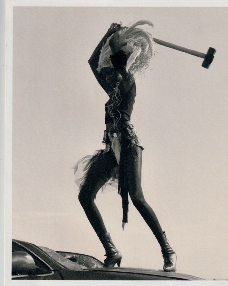 Fashion shot of a woman in costume standing on a car roof with a hammer raised overhead in a black and white outdoor scene.