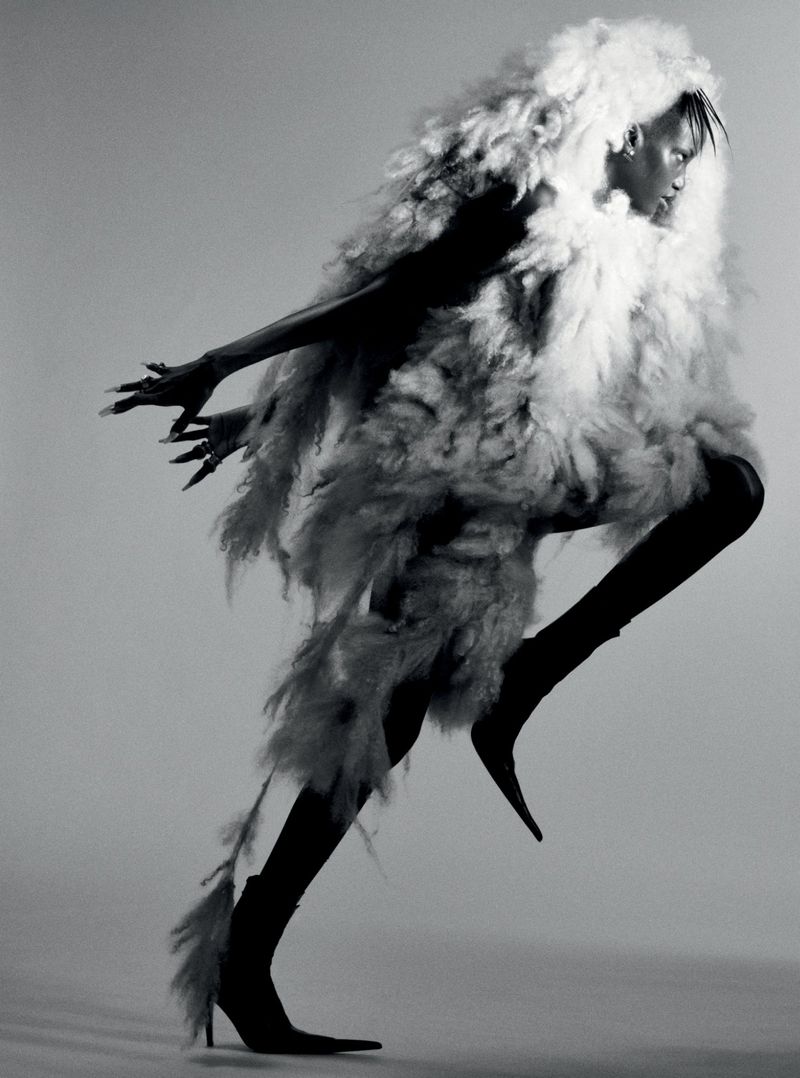 Fashion editorial shot of a Black model in mid-stride, wearing a white feathered coat and high-heeled boots against a grey backdrop.