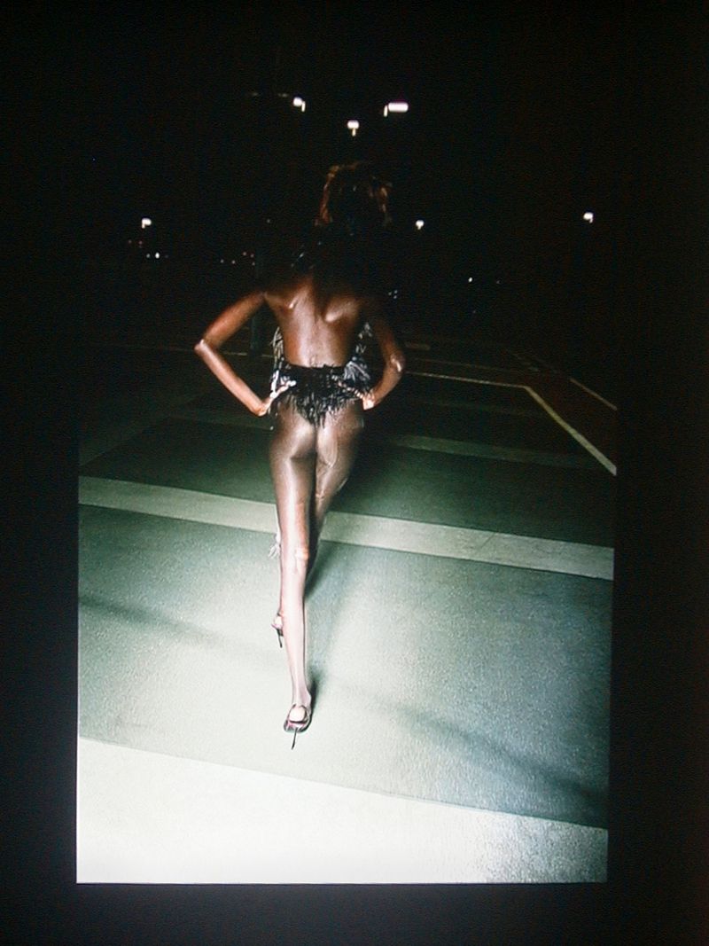 Back view of a slender, dark-skinned woman in a sheer outfit with feathered detailing, walking in high heels outdoors at night.