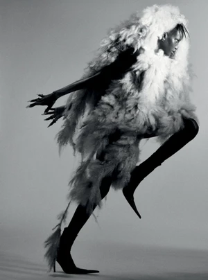 Model Anok Yai in an oversized white feathered coat and black pointed stiletto boots in dynamic motion on a grey studio backdrop.