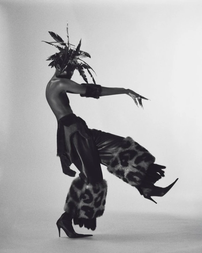 Model Anok Yai posing dynamically in a feather headdress, flared leopard-trimmed trousers, and stiletto heels against a studio backdrop.