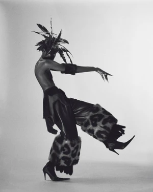 Model Anok Yai posing dynamically in a feather headdress, flared leopard-trimmed trousers, and stiletto heels against a studio backdrop.