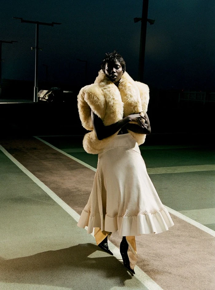 Anok Yai wearing a textured cream bolero shrug and flared skirt standing on a dark outdoor paved court at night.