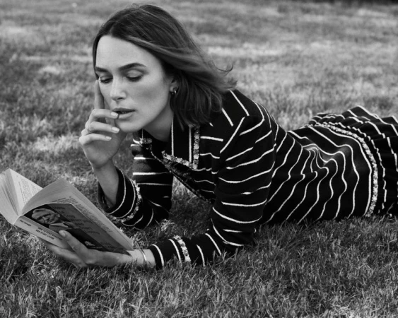 Black and white photo of woman lying in grass reading a book, wearing a striped top, with her finger on her chin.