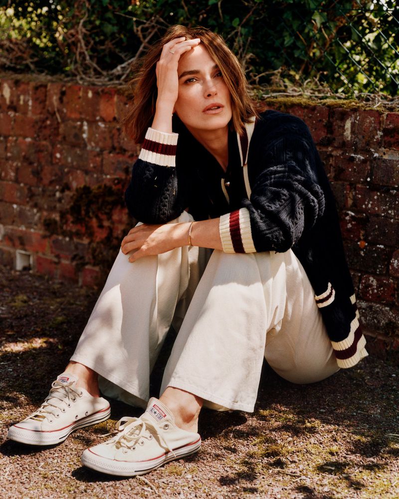 Portrait of Keira Knightley sitting outside with a brick wall and greenery in the background wearing Converse sneakers and a cardigan.