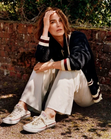 Portrait of Keira Knightley sitting outside with a brick wall and greenery in the background wearing Converse sneakers and a cardigan.