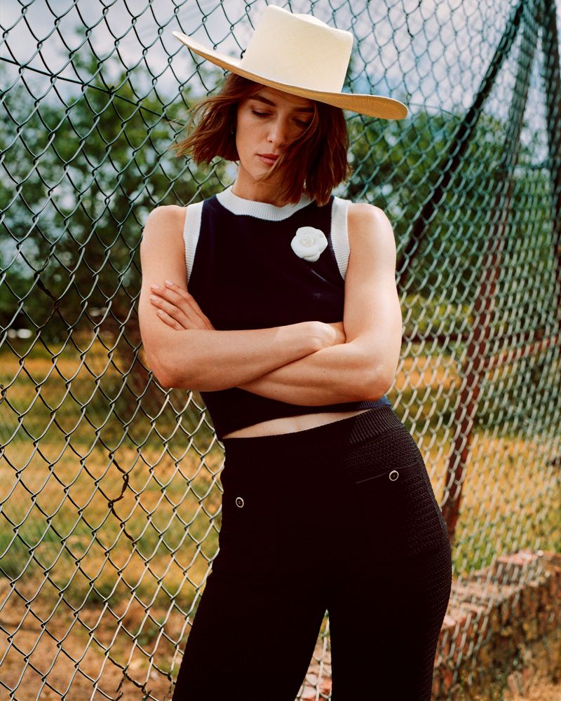 Woman wearing a hat and black knitted outfit stands in front of a chain-link fence with arms crossed in a medium shot.