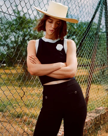 Woman wearing a hat and black knitted outfit stands in front of a chain-link fence with arms crossed in a medium shot.